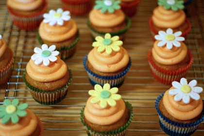 Foto av mange små veganske muffins/cupcakes med oransje krem, pynte med gule, hvite og grønne blomster laget av sukkerfondant.