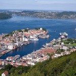 Fotografi av Bergen sett fra Fløien. Flott utsikt over byens havn og husene på hver side, sjøen og lave fjell i det fjerne under en blå himmel.