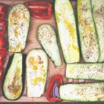 Fotografi av mange halve squash og paprikaer med olje og krydder på seg på en fjøl i lyst tre, sett rett ovenifra.