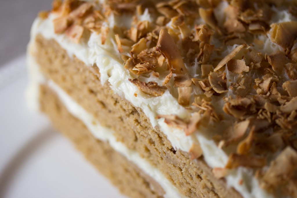 Nærbilde av en kake med lysebrune bunner og hvit krem i midten og på toppen, med lysebrune, ristede kokosflak på toppen.