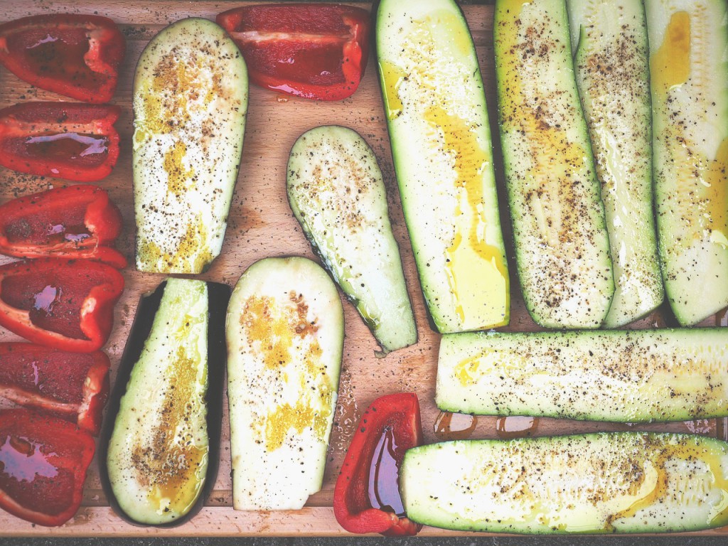 Fotografi av mange halve squash og paprikaer med olje og krydder på seg på en fjøl i lyst tre, sett rett ovenifra.