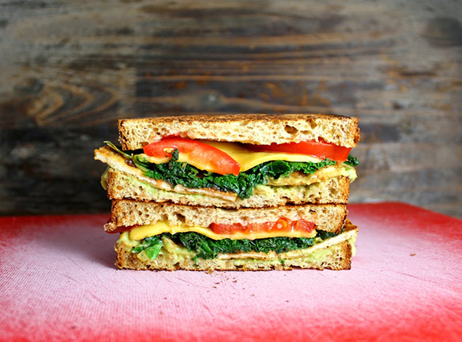 En dobbel sandwich med tofu, veganost, grønnkål og tomat, på en rød-rosa fjøl med en trevegg i bakgrunnen.