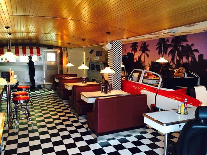 Bilde fra inni en restaurant med amerikansk diner-preg. Sjakkmønster på gulvet, palmer på veggene, røde "båser" med seter, hvite bord.