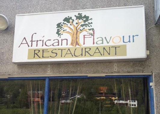 Bilde av et skilt med et tre og "African Flacour Restaurant" på. Under ser man øvre del av et vindu med gardiner.