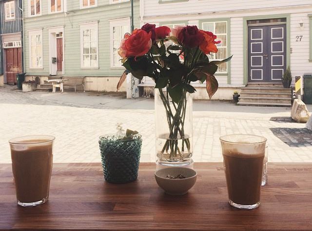 Blomster og to kaffekopper på en bordflate mot et vindu, hvor man ser ut på gata på noen gamle trehus på den andre siden av gata.