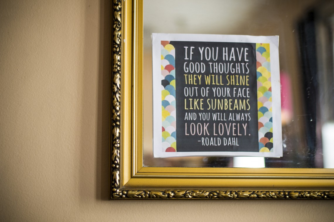Bilde av et hjørne av et speil, med et ark på, med sitat fra Roald Dahl: "If you have good thoughts, they will shine out of you like sunbeams, and you will always look lovely." Hvit tekst på svart bakgrunn, med hvit ramme og farget sirkelmønster rundt. Speilet har gullramme.